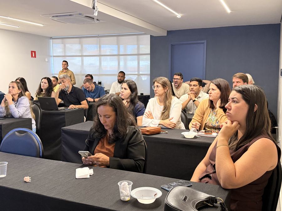 Participantes sentados em mesas de treinamento acompanham a aula em sala com iluminação natural.